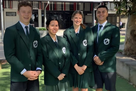 Cuatro personas en togas académicas verdes están frente a un edificio en el campus de la Cavendish Road State High School.