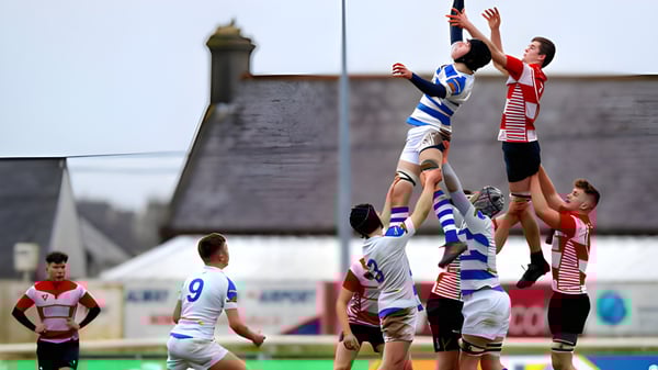 Dos jugadores de rugby luchan por el balón en el aire frente a un edificio histórico en el campus de la C.B.S. Roscommon.