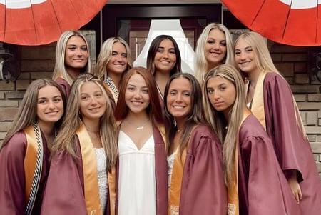 Un grupo de graduadas de la Central Regional High School está de pie en togas de graduación burdeos frente a un edificio de ladrillo con decoraciones rojas y blancas.
