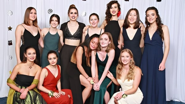 Un grupo de jóvenes estudiantes del Central Southland College posan con diferentes vestidos formales frente a una pared decorada de blanco.