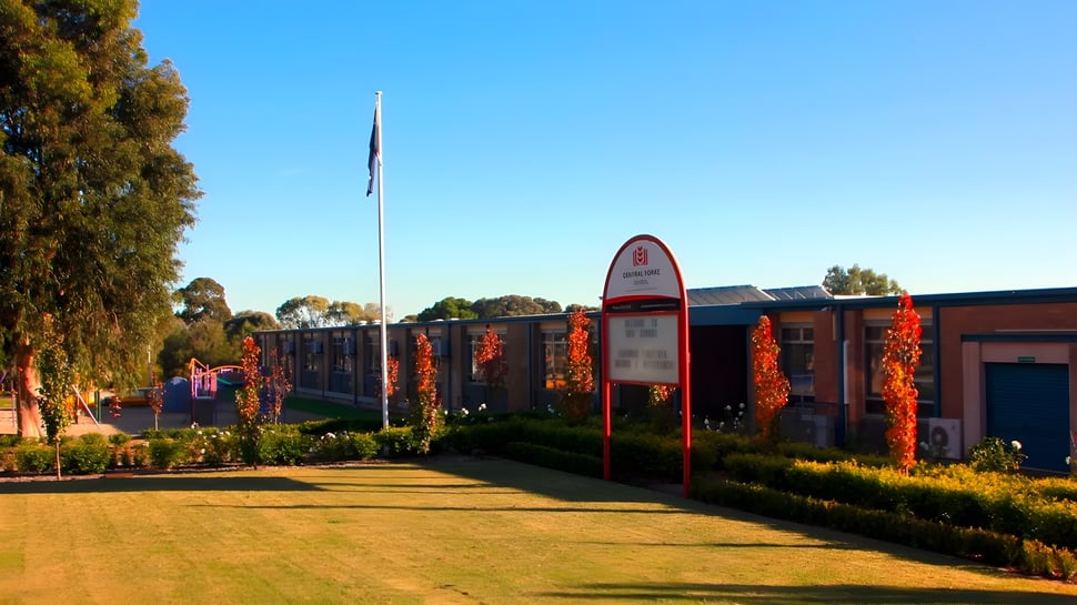 El edificio de ladrillo de la Central Yorke School se encuentra frente a un entorno verde con un alto asta de bandera y un camino pavimentado.