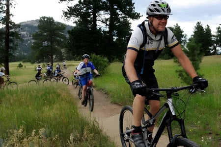 Un grupo de estudiantes del Chancellor State College monta en bicicletas por un camino de campo a través de un prado con pinos.