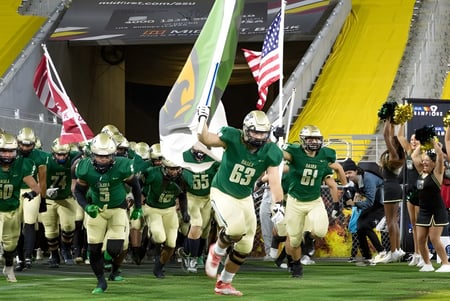 Un grupo de jugadores de fútbol americano en uniformes verdes corre hacia el campo en el Chandler Unified School District.