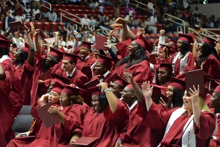 Las graduadas y graduados de las Charlotte-Mecklenburg Schools celebran su ceremonia de graduación en togas rojas frente al escenario y al público.