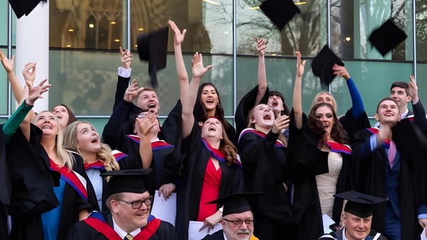 Un grupo de graduados de la Charterhouse School celebra frente a un edificio de vidrio con los brazos en alto por su graduación.