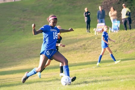 Una jugadora de fútbol de Chapel Hill - Chauncy Hall dribla el balón en el campo con otros jugadores al fondo.