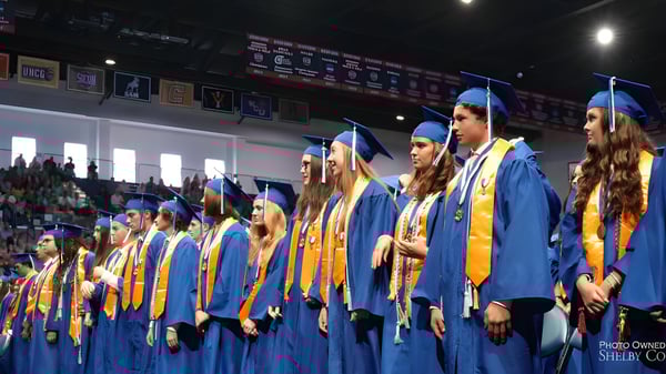 Un grupo de graduados en togas azules y doradas está en el escenario de Chelsea High School durante la ceremonia de graduación.