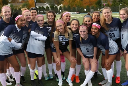 Un grupo de jóvenes atletas de la Cheshire Academy está al aire libre frente a un paisaje otoñal y ríen juntas.
