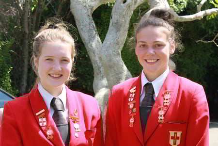 Dos alumnas en uniforme escolar están sonriendo frente a un área boscosa en el campus de la Chilton Saint James School.