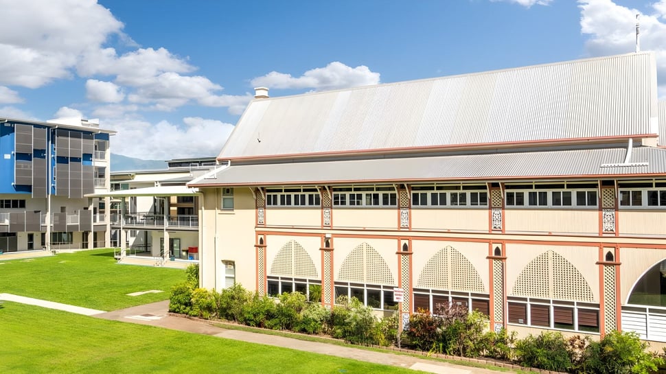 El moderno edificio de varios pisos de la Christ King Secondary School está rodeado de exuberante vegetación bajo un cielo azul con nubes.