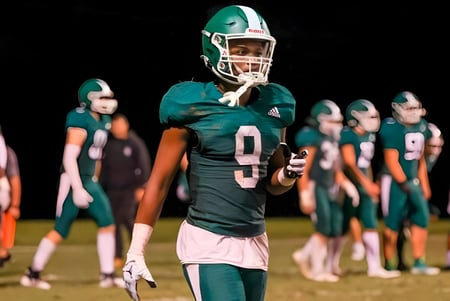 Un jugador de fútbol de la Christ School está en una camiseta verde en el campo con sus compañeros de equipo al fondo.