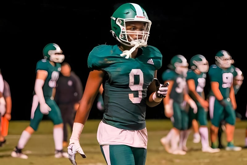 Un jugador de fútbol de la Christ School está en una camiseta verde en el campo con sus compañeros de equipo al fondo.