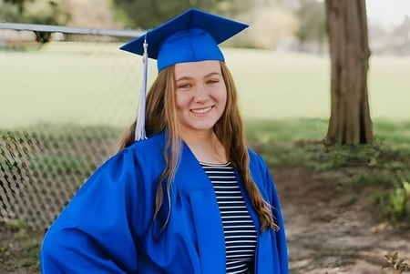 Una joven graduada en una toga de graduación azul está en el campus de la Christian Life School en una pradera con árboles de fondo.