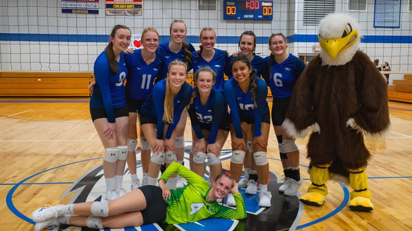 Un grupo de jóvenes jugadoras de voleibol en camisetas azules posan en la cancha de baloncesto de la Christian Life School con la mascota en un disfraz amarillo.