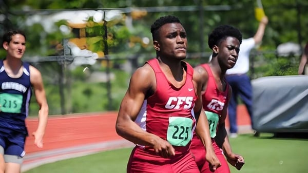 Dos deportistas masculinos corren en la pista de la Church Farm School con un campo cubierto de hierba y árboles al fondo.