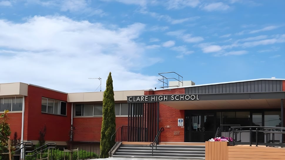 El edificio de ladrillo rojo de Clare High School está rodeado de árboles y se encuentra bajo un cielo azul nublado.
