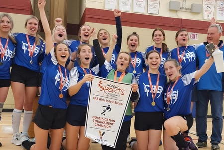 Las estudiantes de la Clarenville High School celebran su victoria con medallas y trofeo frente a una pared pintada de rojo.