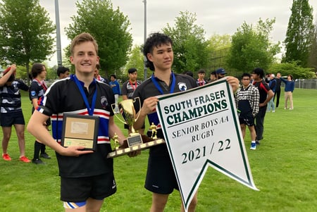 Dos estudiantes de la Clayton Heights Secondary School sostienen un trofeo y una pancarta de campeonato al aire libre frente a una multitud.