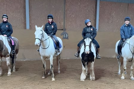 Estudiantes de la Colaiste Chiarain Athlone montan a caballo en un salón con suelo de tierra marrón y paredes de madera.