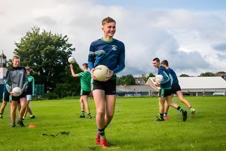Un grupo de jóvenes atletas juega en el campo deportivo de Coláiste Chríost Rí frente a árboles y edificios.