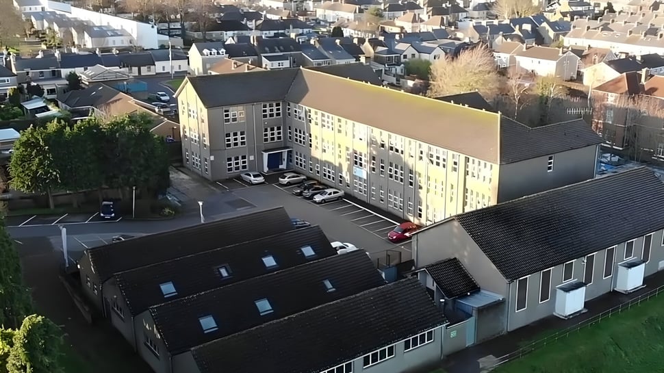 El edificio principal de varios pisos de la Coláiste Éamann Rís se encuentra en primer plano frente a un paisaje urbano.