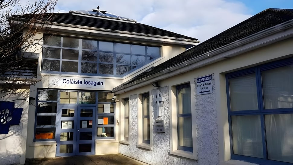 El edificio de Coláiste Íosagáin es de piedra con un techo inclinado y grandes ventanas bajo un cielo nublado rodeado de árboles desnudos.