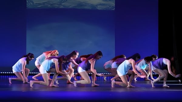 Un grupo de bailarinas y bailarines presenta una actuación de baile en el escenario de Coláiste Mhuire (Ballygar) con trajes coloridos sobre un fondo azul.