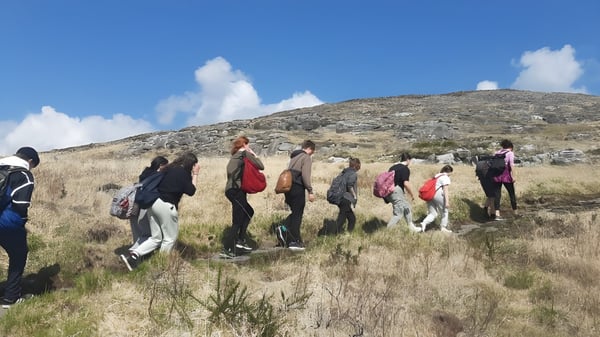 Un grupo de estudiantes de Coláiste Muire Crosshaven camina por un sendero cubierto de hierba en un paisaje montañoso bajo un cielo azul con nubes.