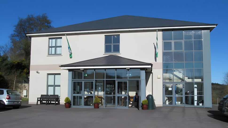 El moderno edificio de dos pisos con fachada de vidrio de Coláiste Muire Crosshaven está rodeado de un estacionamiento pavimentado y árboles.