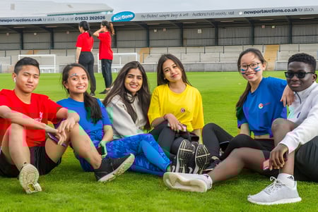 Un grupo de alumnas y alumnos está sentado en un prado frente a un estadio deportivo en el campus de Coláiste Muire Crosshaven.