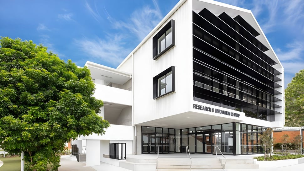 El moderno edificio de varios pisos de la Coláiste Naomh Cormac está rodeado de vegetación verde bajo un cielo azul.