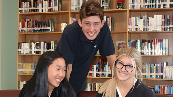 Tres jóvenes adultos están juntos en la biblioteca del campus del Collège Notre-Dame entre estanterías de libros.