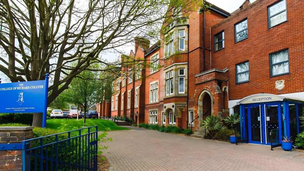 El edificio de ladrillo con ventanas curvas del College of Richard Collyer se encuentra junto a un camino pavimentado y está rodeado de árboles.