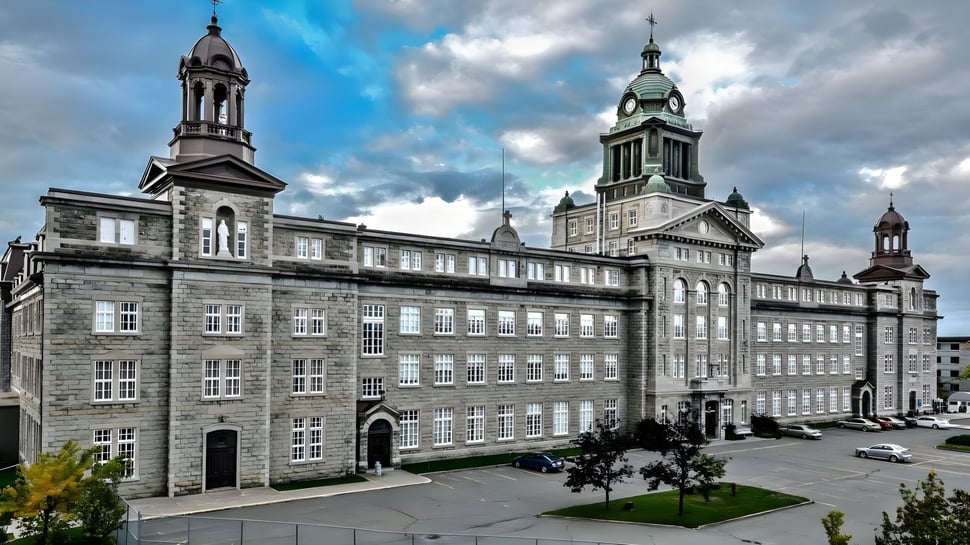 El histórico edificio principal del Collège de Sainte-Anne-de-la-Pocatière con torres y cúpulas rodeado de un patio pavimentado y árboles verdes.
