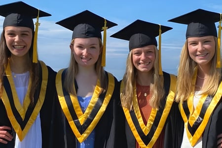 Cuatro estudiantes con ropa de graduación están de pie bajo un cielo azul en el campus del Collingwood Collegiate Institute.