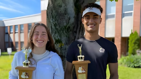 Dos personas están frente a un árbol en el campus de la Colonel Gray High School sosteniendo trofeos en la mano.