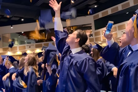 Un grupo de graduados de la Colorado Springs Christian School celebra en túnicas azules y con los brazos levantados en un interior festivamente iluminado.