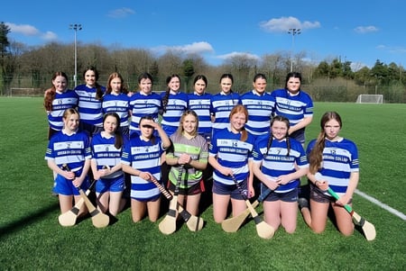 Un grupo de estudiantes del Comeragh College posan juntos en camisetas a rayas azul y blanco en un campo de fútbol frente a un bosque y un cielo azul.
