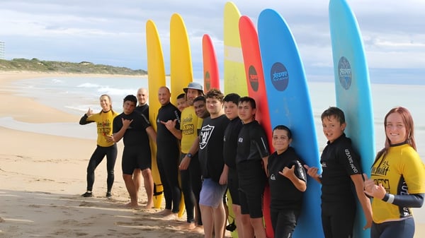Estudiantes del Coodanup College están en trajes de neopreno en la playa con coloridas tablas de surf frente a un paisaje costero.