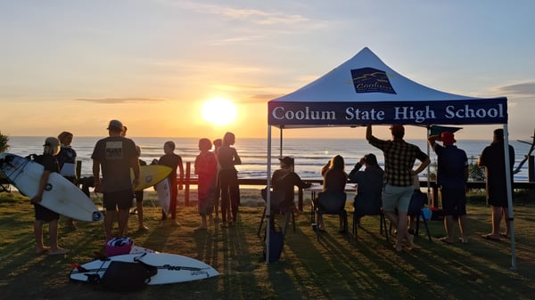 Un grupo de estudiantes está frente al banner de la Coolum State High School en la playa durante el atardecer.