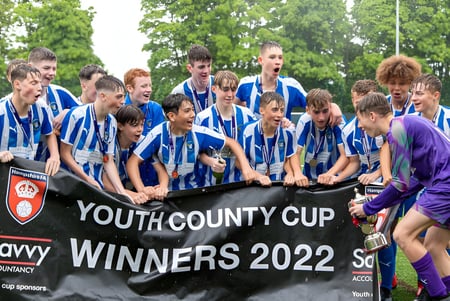 Un grupo de estudiantes de la Corfe Hills School está en un campo de césped alrededor de un banner por la victoria en la Youth County Cup 2022.