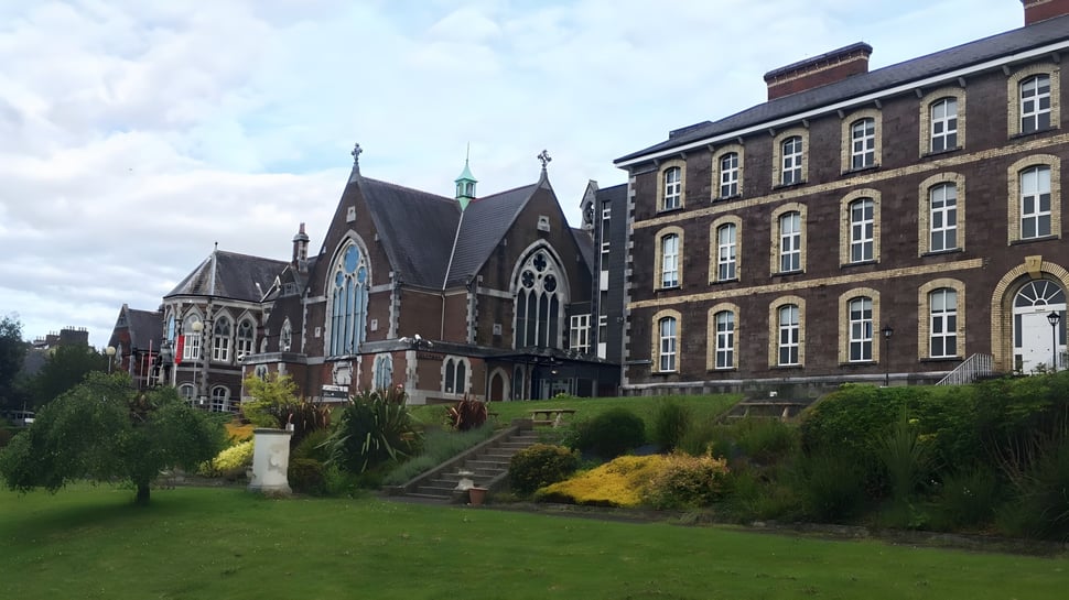 El histórico edificio de la Cork Educate Together Secondary School con un jardín bien cuidado en primer plano bajo un cielo nublado.