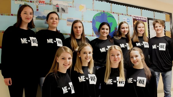 Un grupo de estudiantes con camisetas negras de ME + WE está frente a una pared cubierta de materiales de aprendizaje en la Corner Brook Regional High School.