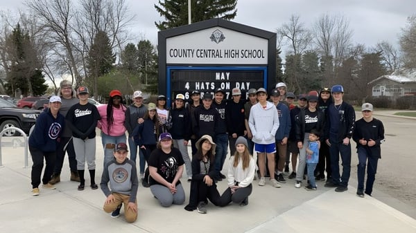 Un grupo de estudiantes y personal se encuentra frente al letrero de la County Central High School con un bosque y edificios al fondo.