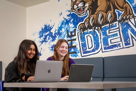 Dos estudiantes de la Covenant Christian School están sentadas en una mesa y trabajan en laptops frente a un mural de tigre.