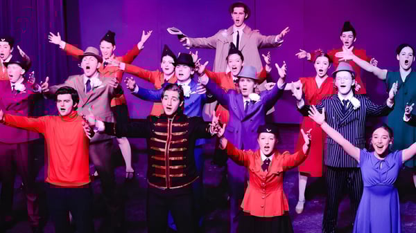 Estudiantes de la Covenant Day School realizan una animada actuación en el escenario con coloridos disfraces y los brazos en alto.