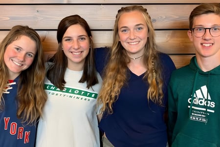 Cuatro estudiantes de la Covenant Day School posan sonriendo frente a un panel de madera.