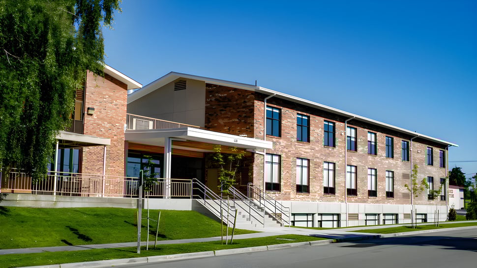 El moderno edificio de ladrillo de la Covenant High School con grandes ventanas se encuentra frente a una pradera bajo un cielo azul.
