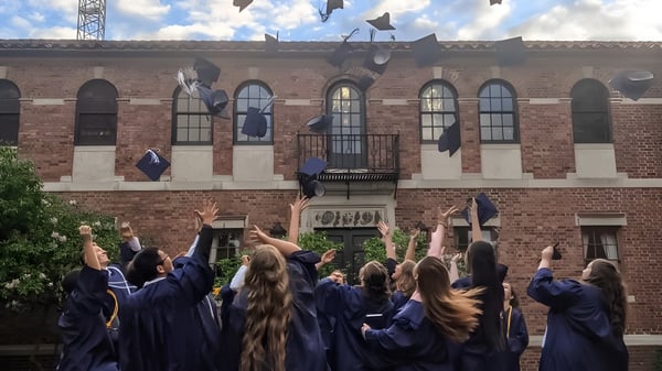 Las graduadas y graduados de la Covenant High School en togas azules reunidos frente a un edificio de ladrillo con torre.