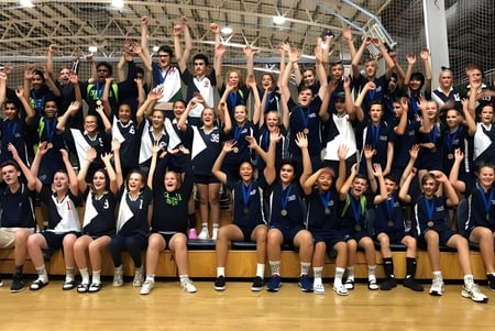 Un grupo de estudiantes de la Craigslea State High School posan juntos en el gimnasio con los brazos en alto.
